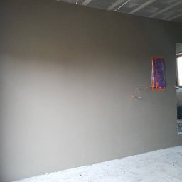 Renova Lublin - Świeżo nałożona szara powłoka na ścianie w surowym wnętrzu, zabezpieczone kable elektryczne, widoczny betonowy sufit i fragment okna w tle.