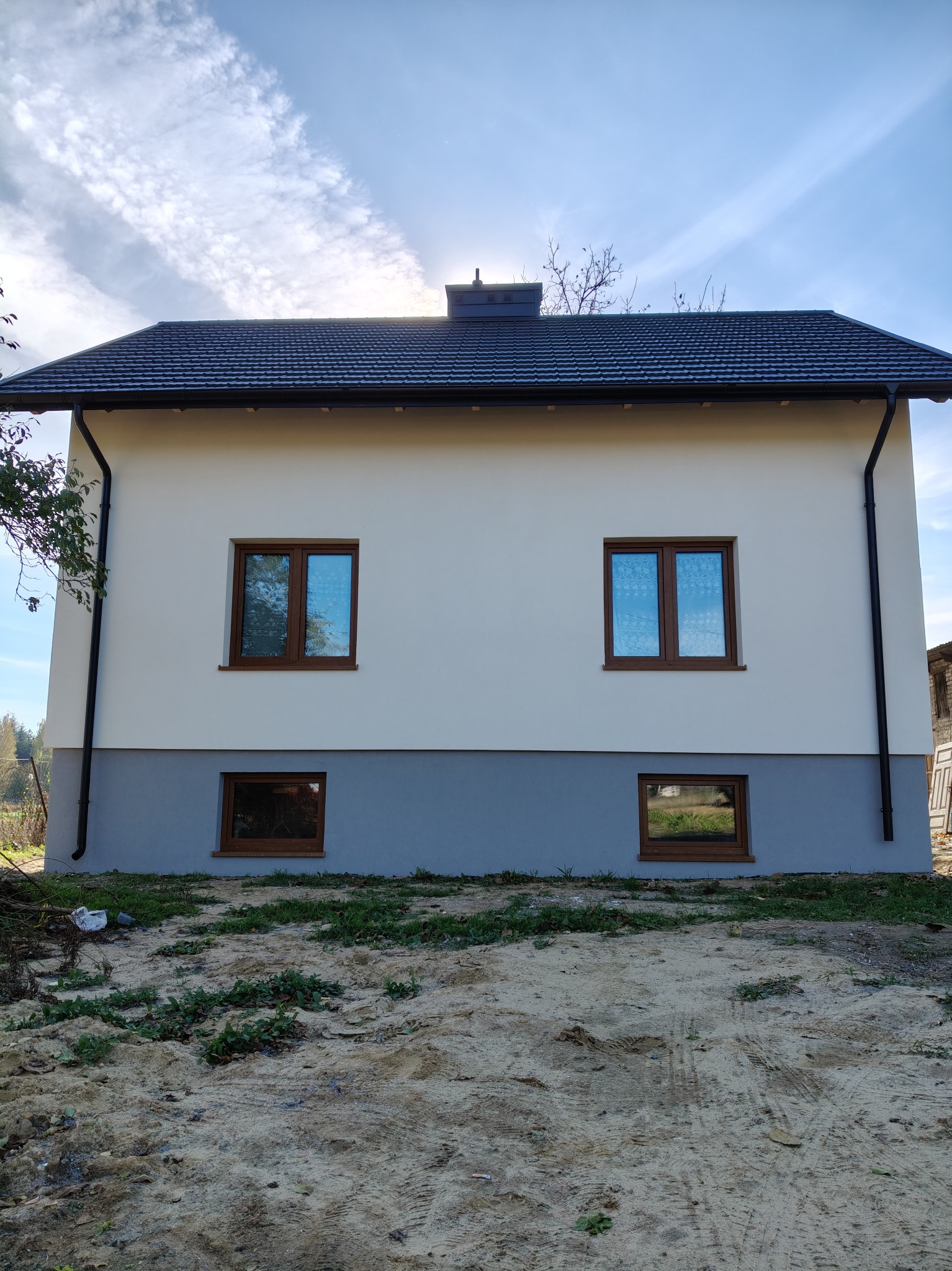 Dwukondygnacyjny budynek z nową, jasną elewacją, brązowymi oknami i grafitowym cokołem, widoczny w słoneczny dzień z perspektywy ziemi.