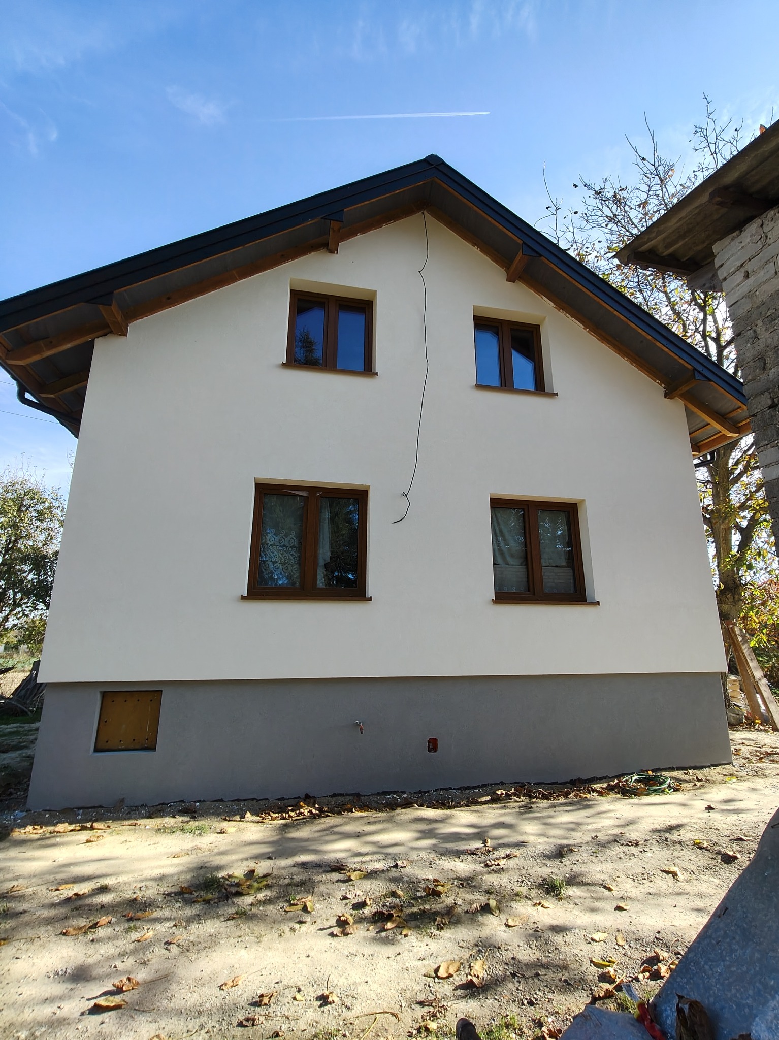 Dwupiętrowy dom z oknami w brązowych ramach, świeżo otynkowany na biało, z szarym cokołem, widoczny z zewnątrz w słoneczny dzień. Na ścianie widoczna pionowa rysa.