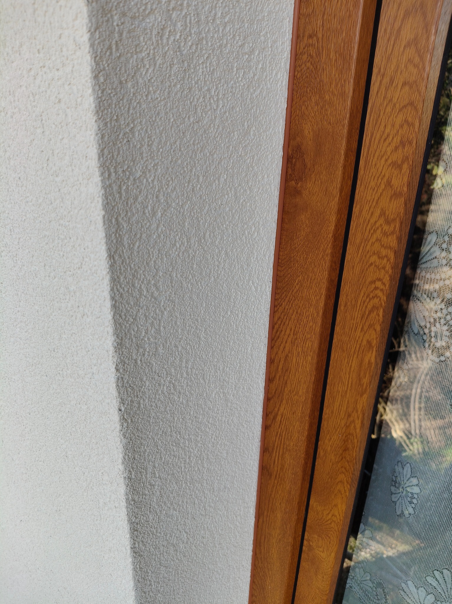 Fragment ściany z gładzią strukturalną obok brązowej ramy okiennej z widoczną siatką przeciw owadom.