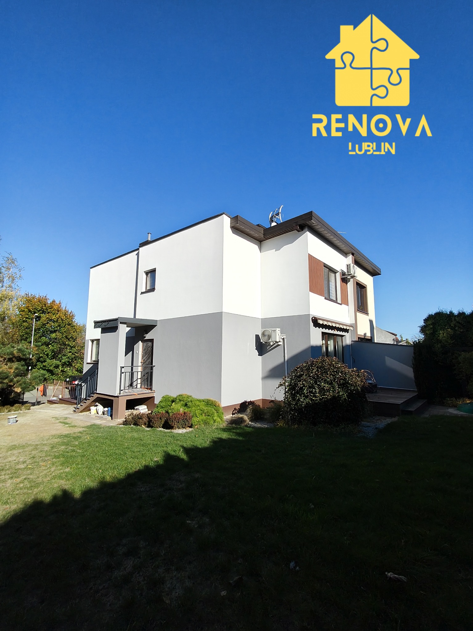 Dwukondygnacyjny dom z szarą dolną i białą górną częścią elewacji, widoczny z trawnika w słoneczny dzień, z logo firmy na niebie.