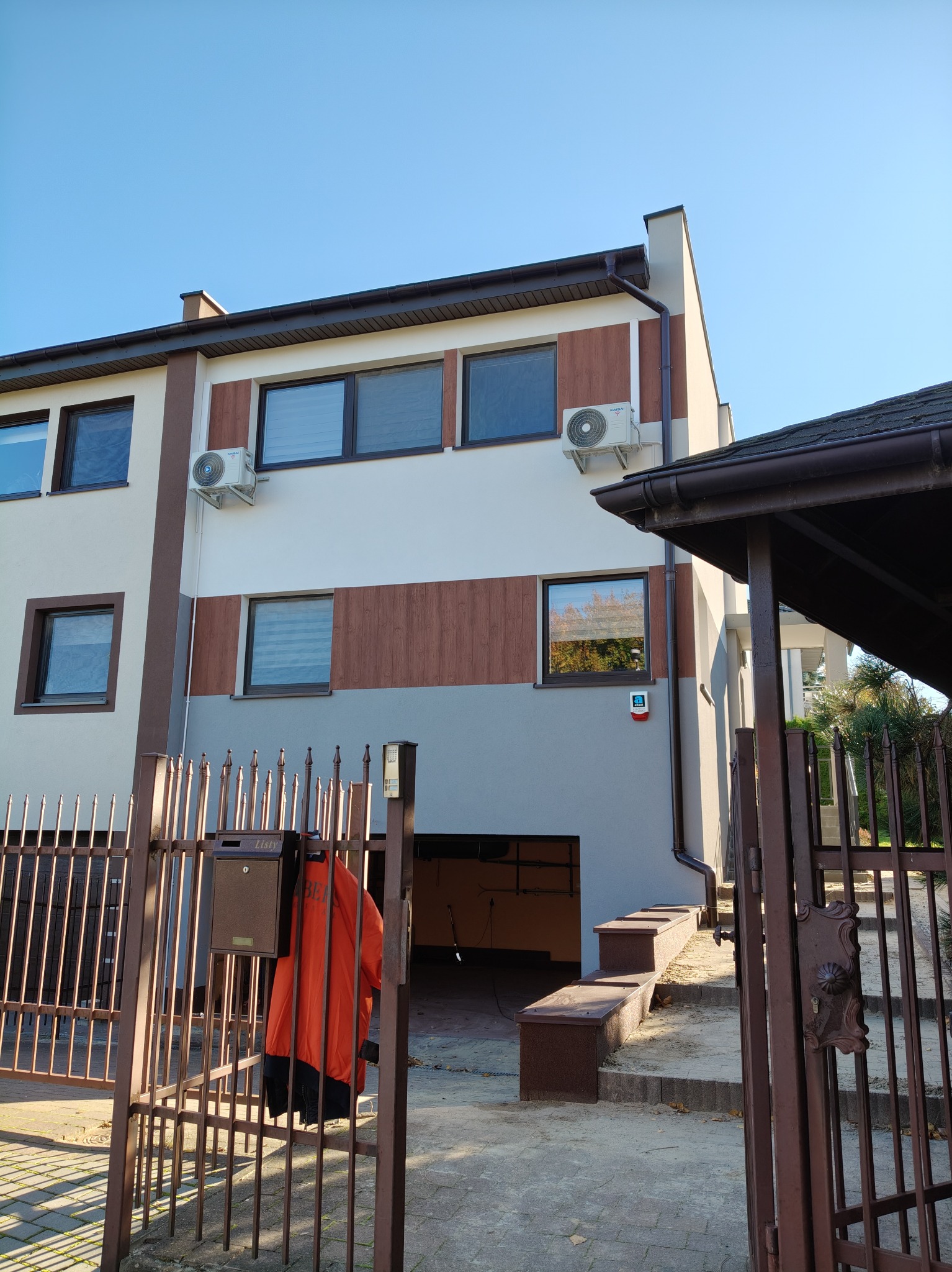 Dwukondygnacyjny dom z szaro-białą elewacją, brązowymi akcentami i dwoma klimatyzatorami na ścianie, widziany zza metalowego ogrodzenia z wiszącą pomarańczową kurtką.