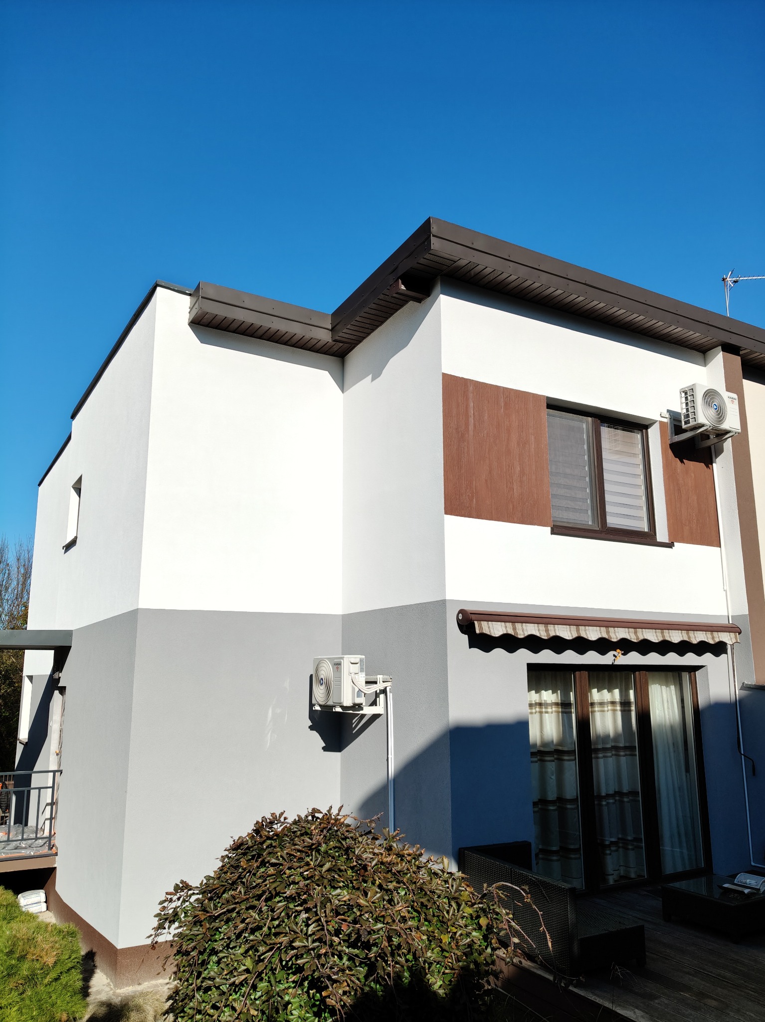 Dwukondygnacyjny dom z nową elewacją w odcieniach bieli i szarości, z brązowymi akcentami i klimatyzatorami na ścianie, sfotografowany w słoneczny dzień.