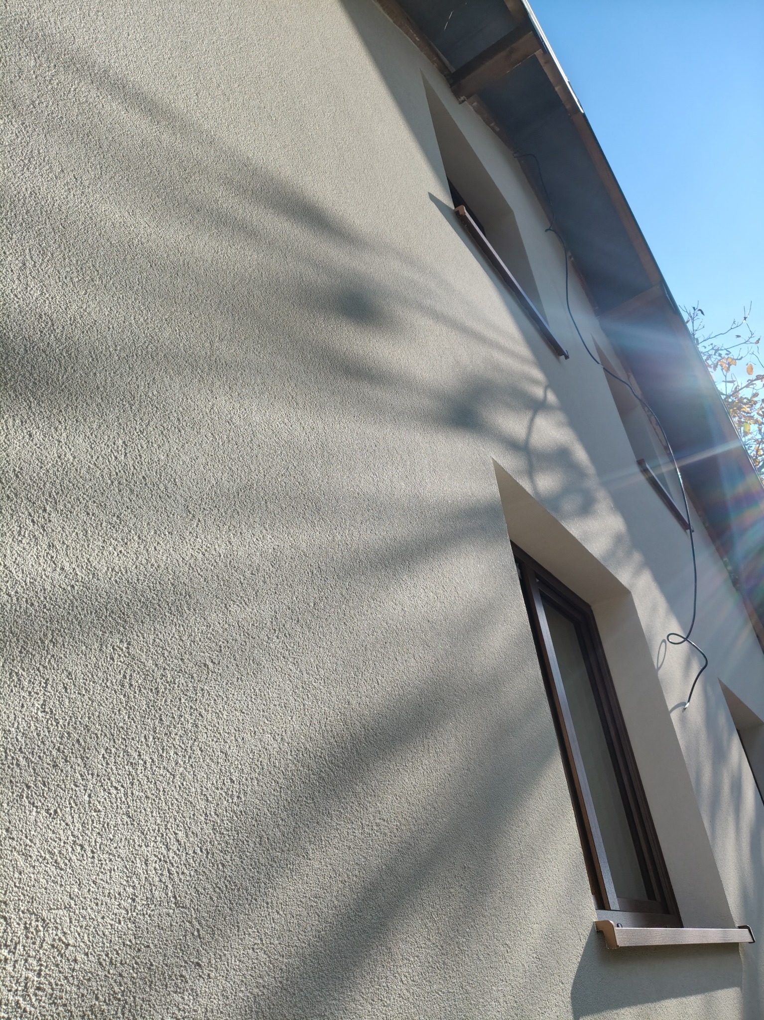 Jasna elewacja budynku z widoczną fakturą, oświetlona słońcem, z oknami w ciemnych ramach i widocznymi cieniami drzew.