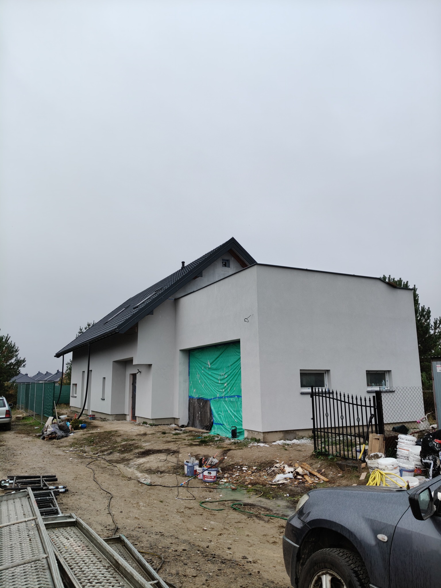 Nowoczesny dom jednorodzinny z szarą elewacją i ciemnym dachem, częściowo zasłonięty zieloną plandeką podczas prac wykończeniowych, widoczne rusztowania i materiały budowlane na posesji w pochmurny...