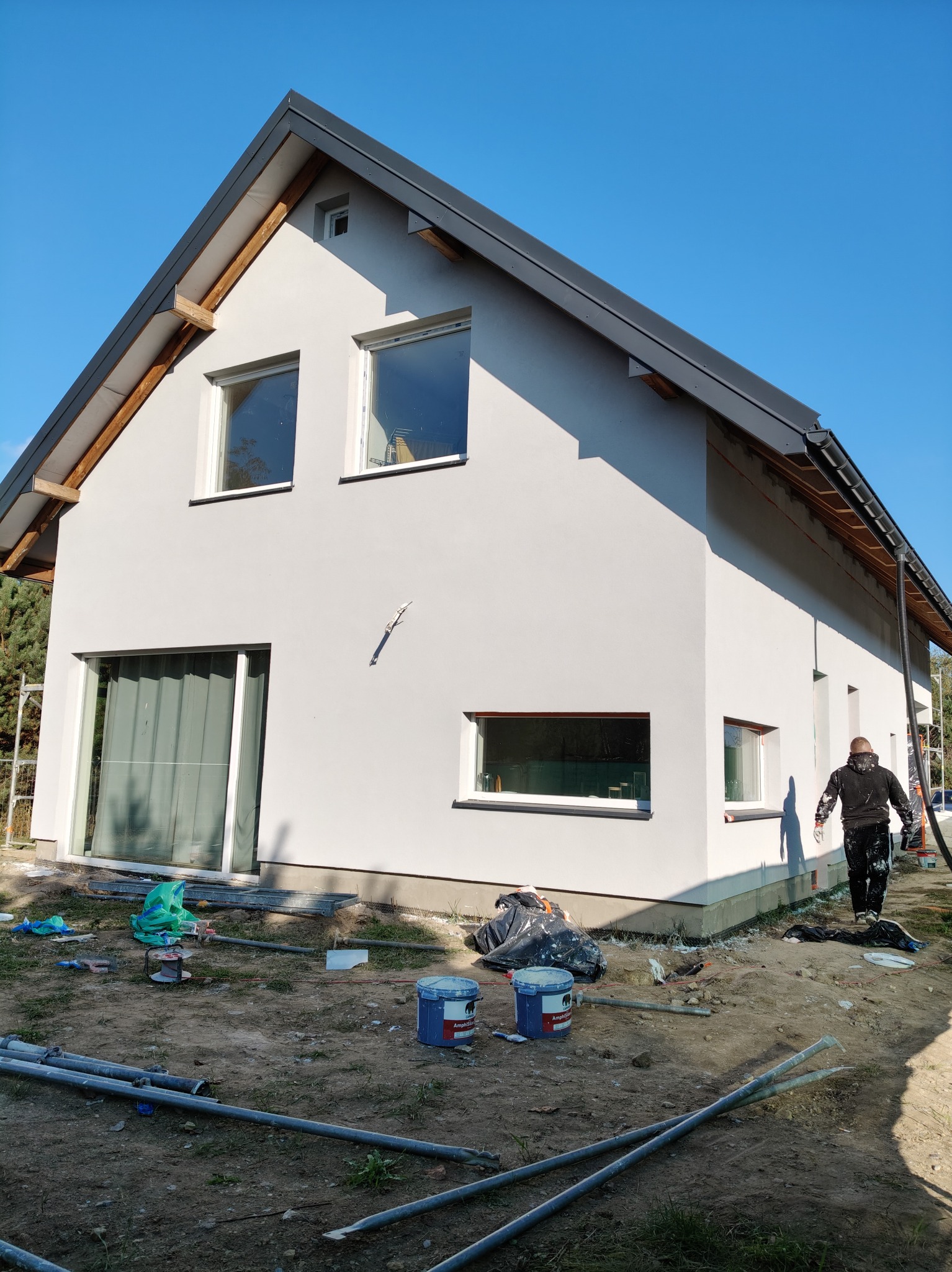 Świeżo otynkowany dom jednorodzinny w jasnych barwach, z widocznymi śladami prac wykończeniowych wokół budynku i przygotowanym sprzętem do dalszych prac na elewacji, na tle błękitnego nieba.