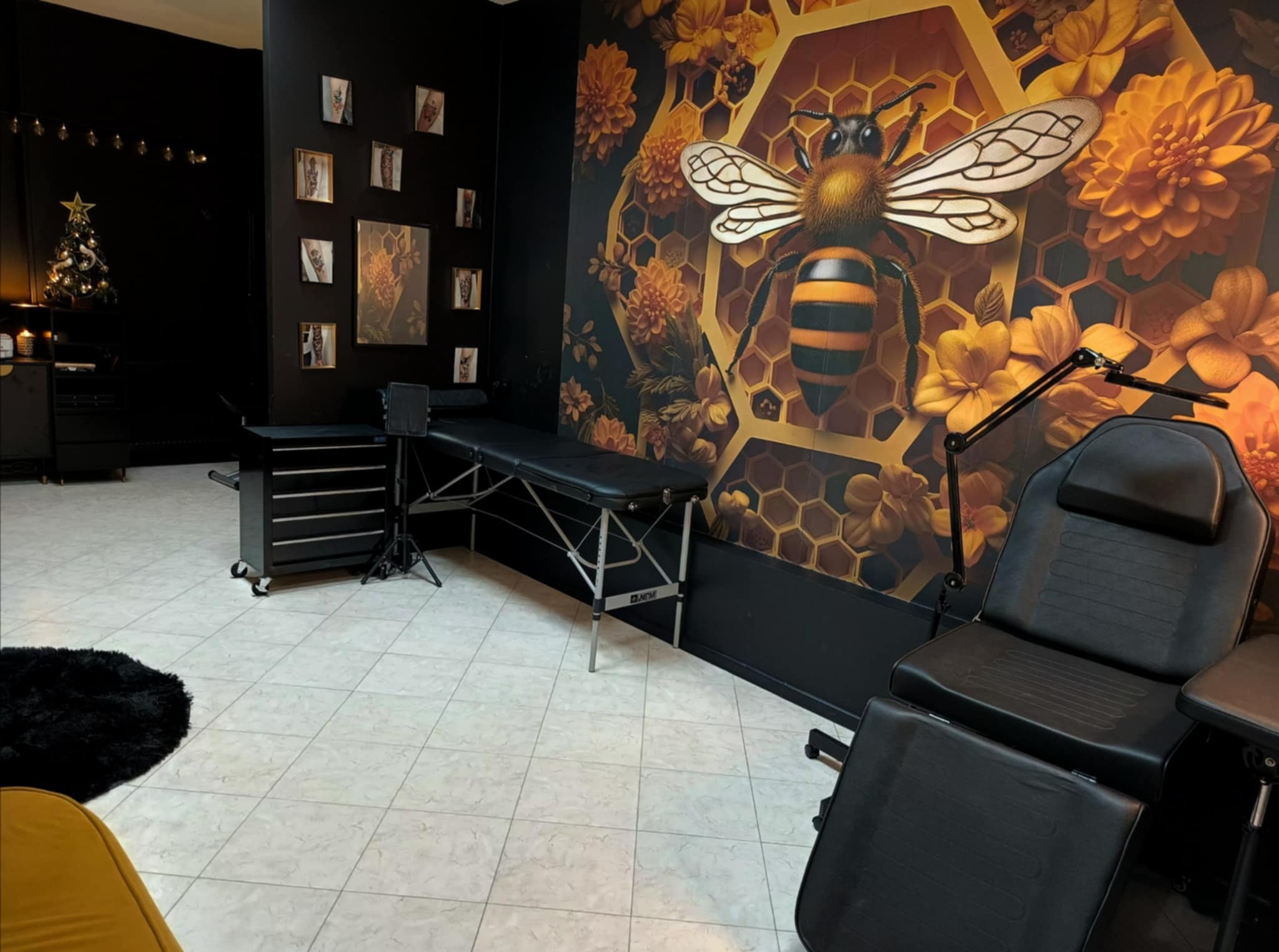 Wnętrze salonu tatuażu z czarnymi ścianami, wzorem plastra miodu i motywem pszczoły na ścianie, czarnym fotelem do tatuażu, w tle choinka i ramki ze wzorami tatuaży.