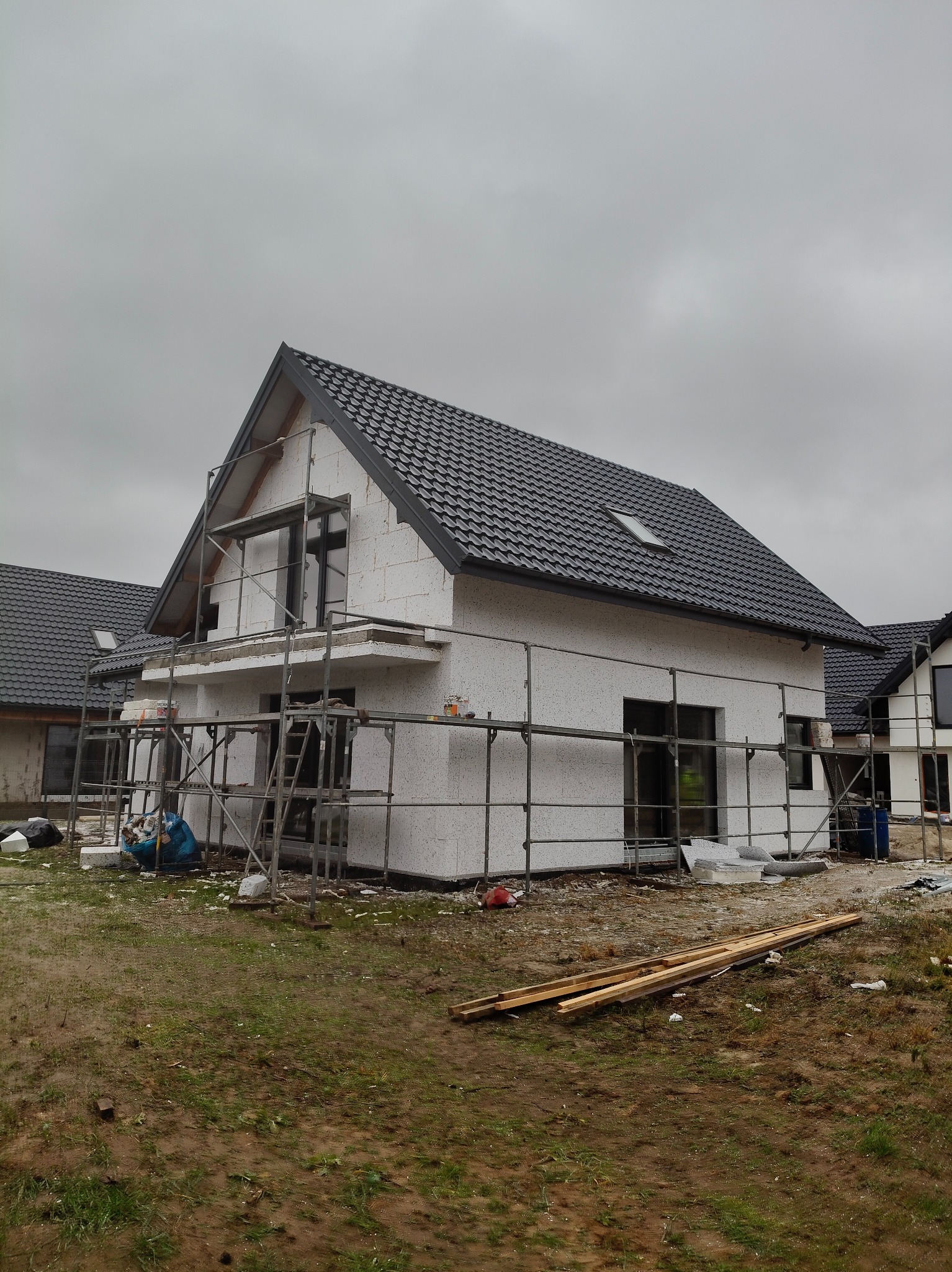 Dom jednorodzinny w trakcie prac elewacyjnych, widoczne rusztowania, biała fasada z szarym dachem i elementami okiennymi, pochmurne niebo w tle.