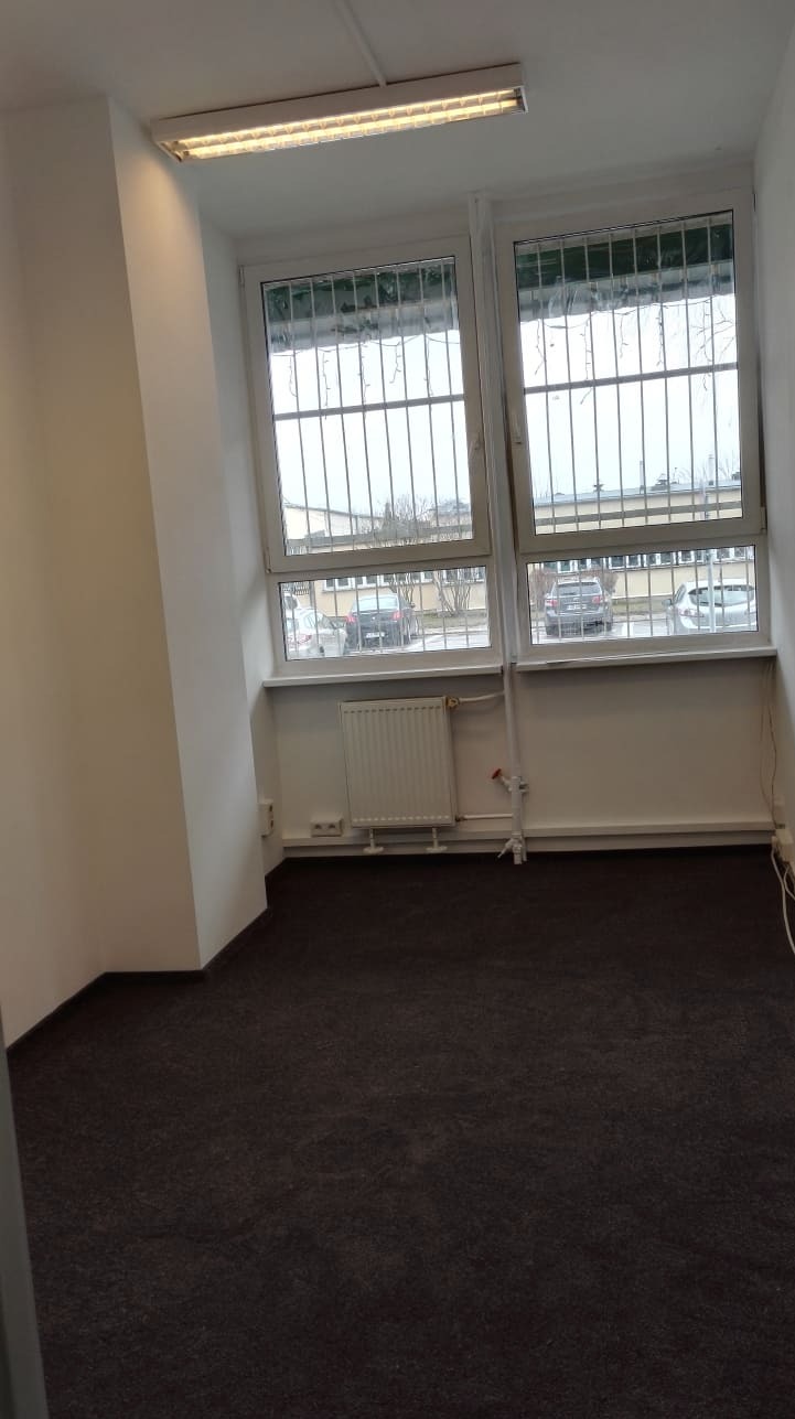 Puste pomieszczenie biurowe z ciemną wykładziną, białymi ścianami, grzejnikiem i oknami z kratami, widok na parking.