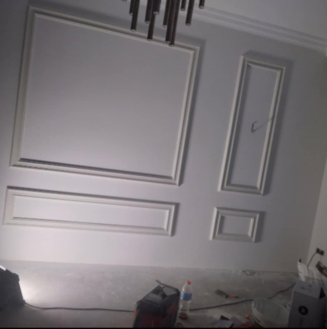 Ściana z dekoracyjnymi, białymi ramami o różnych kształtach i rozmiarach, w trakcie prac wykończeniowych, widoczne narzędzia i butelka wody na podłodze.