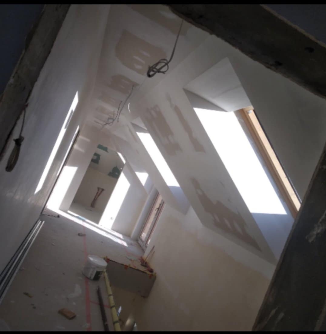 Poddasze w trakcie prac wykończeniowych, widoczne okna dachowe, ściany z nałożoną gładzią szpachlową przed szlifowaniem, kabel zwisający z sufitu, oraz narzędzia malarskie na podłodze.