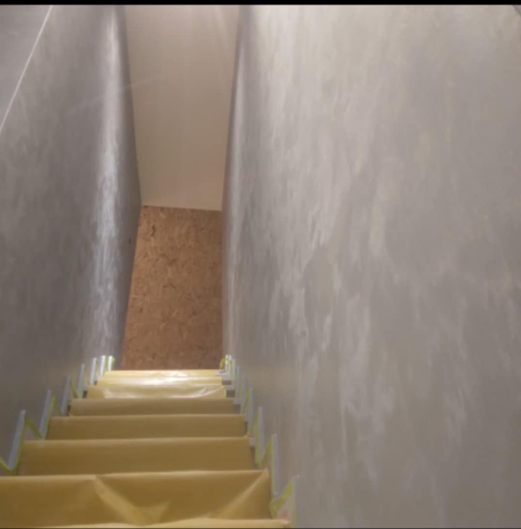 Klatka schodowa w trakcie prac wykończeniowych; schody zabezpieczone folią ochronną, ściany pokryte jasnoszarą, dekoracyjną strukturą imitującą marmur.