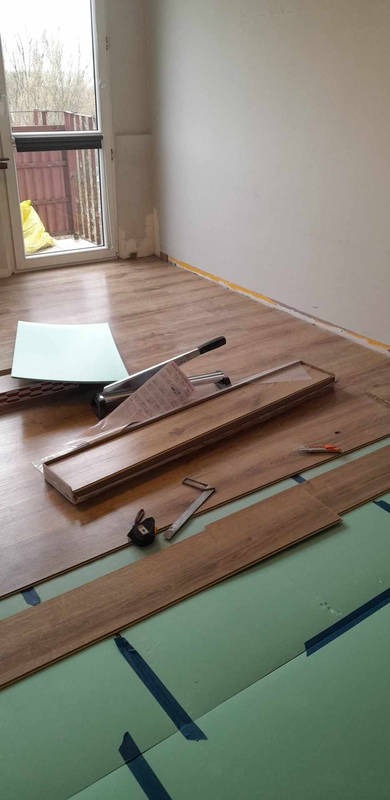 Fragment pokoju w trakcie montażu podłogi z paneli drewnopodobnych, widoczne podłoże z zielonej pianki mocowane niebieską taśmą, narzędzia do cięcia i montażu paneli oraz przycięte deski.