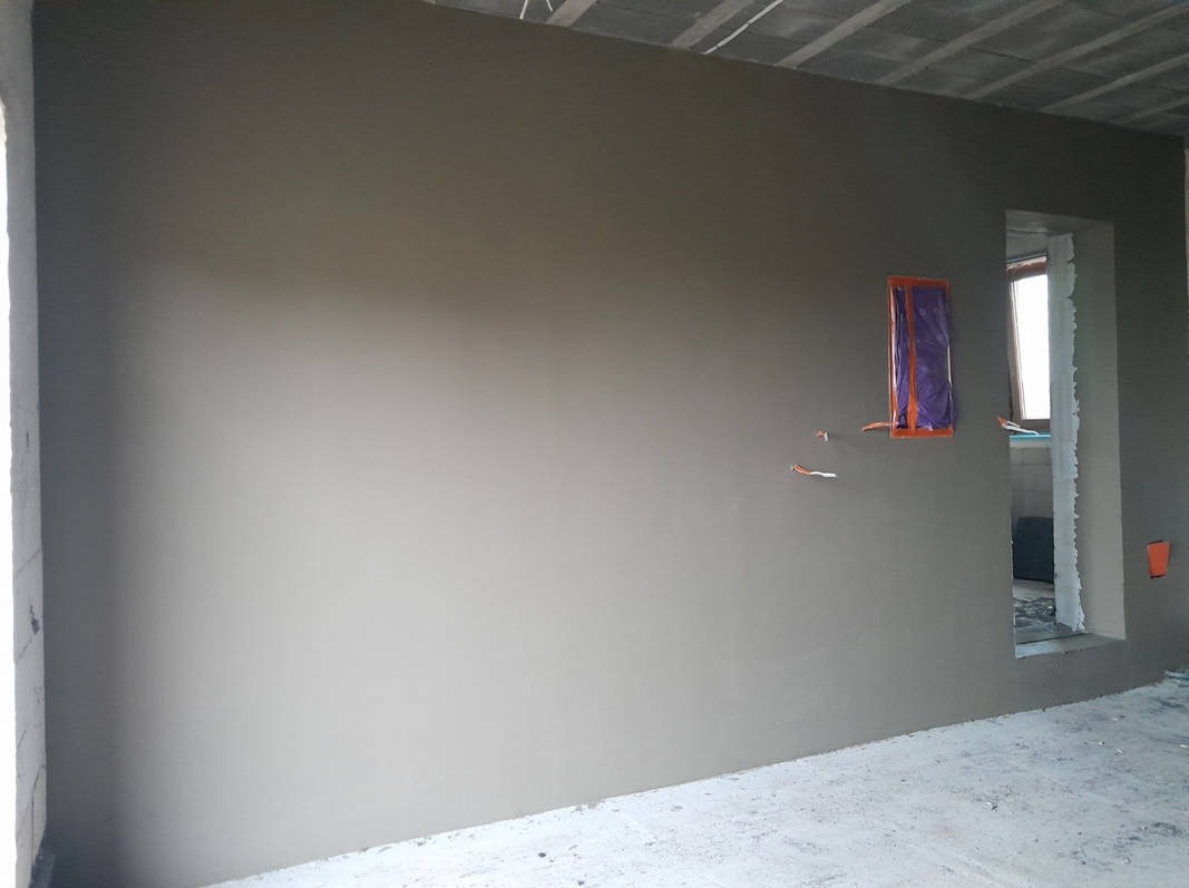 Świeżo nałożona szara powłoka na ścianie w surowym wnętrzu, zabezpieczone kable elektryczne, widoczny betonowy sufit i fragment okna w tle.