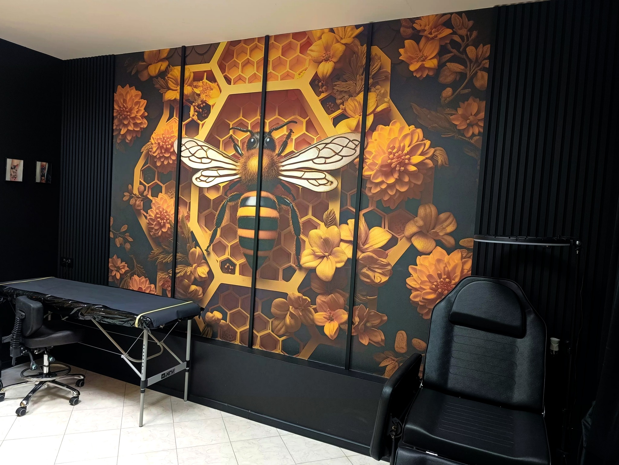 Wnętrze gabinetu z czarnymi ścianami i dekoracyjnym panelem ściennym przedstawiającym pszczołę na tle plastra miodu i kwiatów; widoczne czarne krzesło i stół zabiegowy.