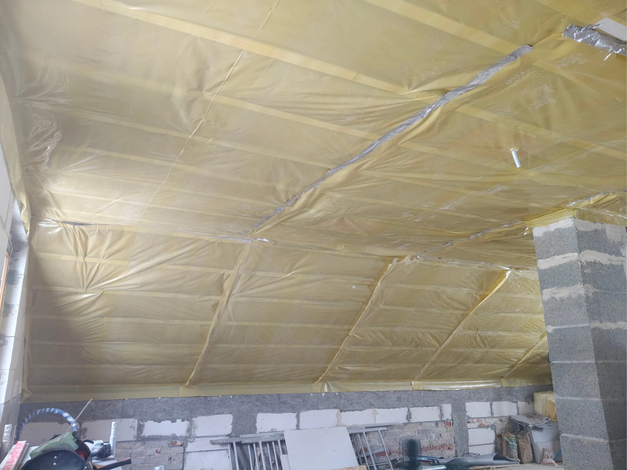 Izolacja poddasza folią paroizolacyjną, widoczne drewniane krokwie i fragment ściany z betonu komórkowego z widocznymi śladami zaprawy.