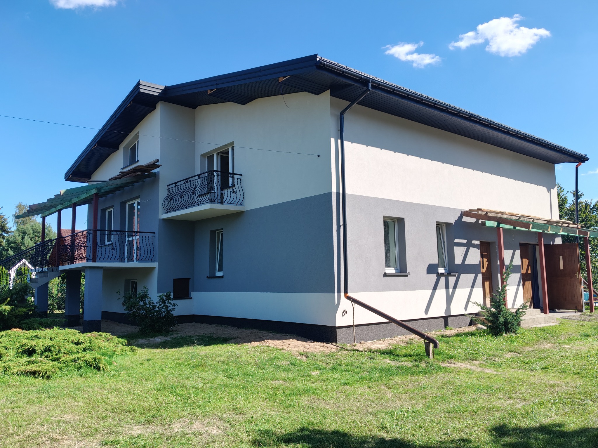Dwukondygnacyjny dom z ocieploną i pomalowaną elewacją w odcieniach szarości i bieli, z balkonami z kutą balustradą, widoczny z perspektywy trawnika w słoneczny dzień.
