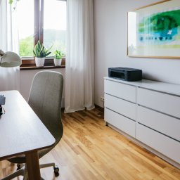 Chodaccy Dom i Wnętrze - Jasne biuro domowe z drewnianym biurkiem, szarą tapicerowaną krzesłem, białą komodą z drukarką, lampką biurkową i abstrakcyjnym obrazem na ścianie.