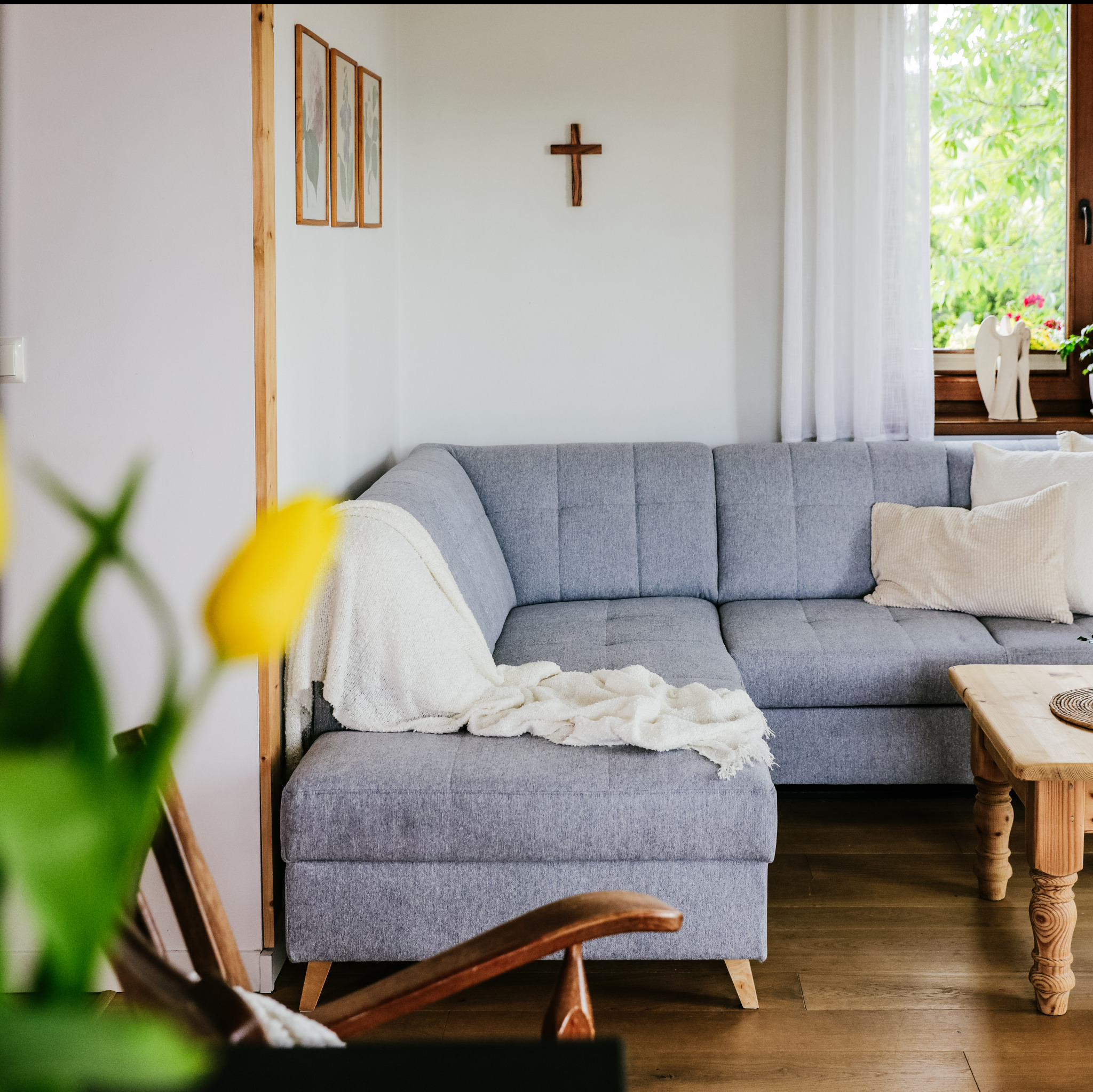 Jasnoszary narożnik z pledem, drewniany stolik kawowy i krzesło na pierwszym planie, widok na okno z zielenią za firanką i drewniany krzyż na ścianie.