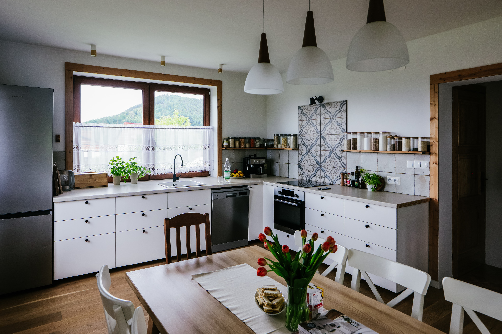 Jasna kuchnia z białymi szafkami i drewnianymi akcentami, widok na zieleń za oknem, stół z tulipanami i ciasteczkami na pierwszym planie.