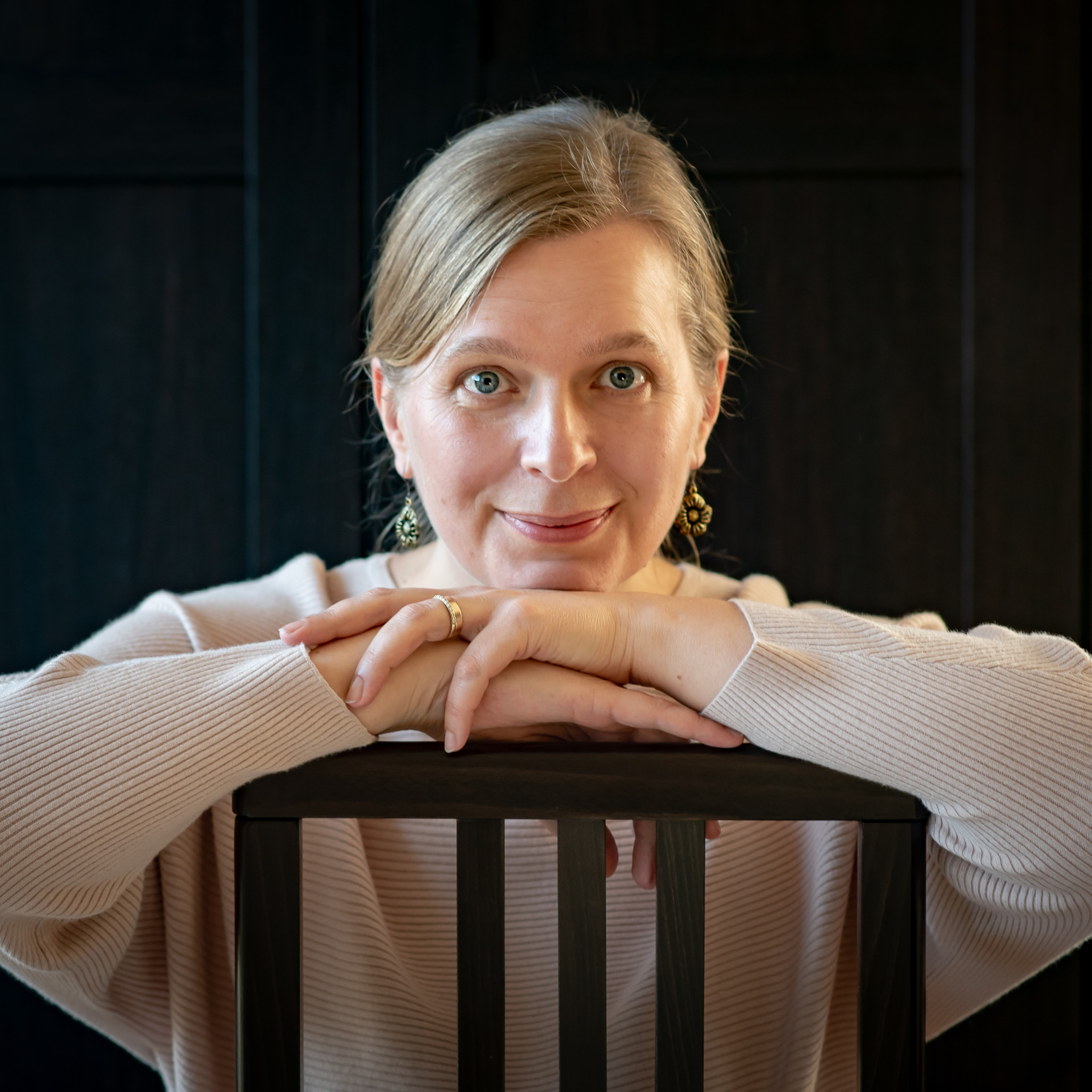Portret kobiety z blond włosami i niebieskimi oczami, opierającej się o czarne krzesło z drewnianymi szczebelkami, w tle ciemna ściana.
