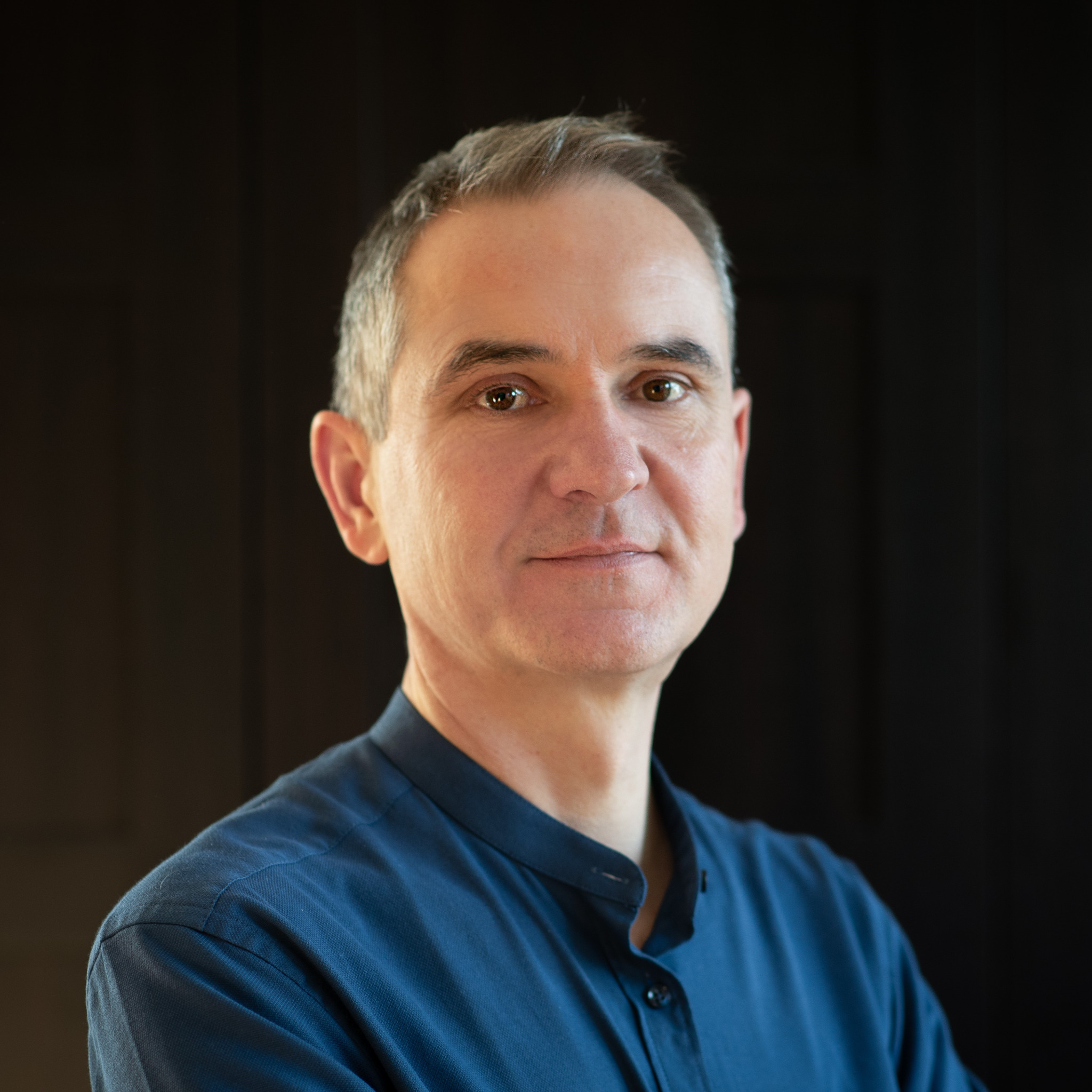 Portret mężczyzny z siwiejącymi włosami, ubranego w niebieską koszulę na tle ciemnego, jednolitego tła.