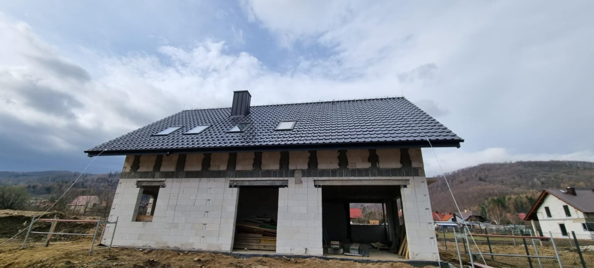 Dom w stanie surowym zamkniętym z czarną dachówką i oknami dachowymi, widoczne bloczki betonowe i otwory okienne, w tle zalesione wzgórza i pochmurne niebo.