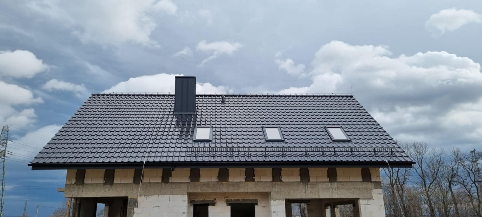 Dom w stanie surowym zamkniętym z grafitową dachówką, trzema oknami dachowymi i kominem na tle pochmurnego nieba.