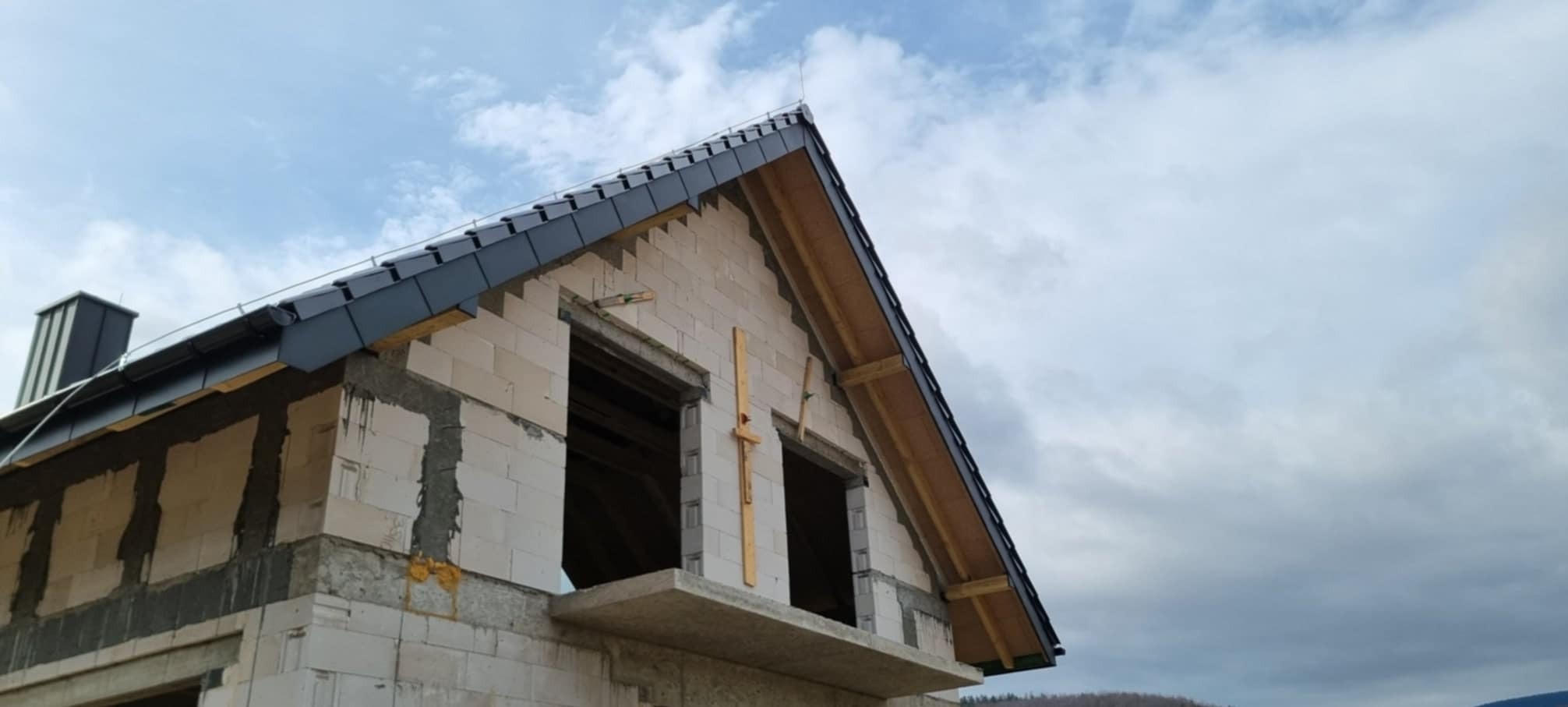 Niedokończony dom z białego pustaka z czarnym dachem z blachy na tle pochmurnego nieba, widoczne otwory okienne z drewnianymi stemplami.