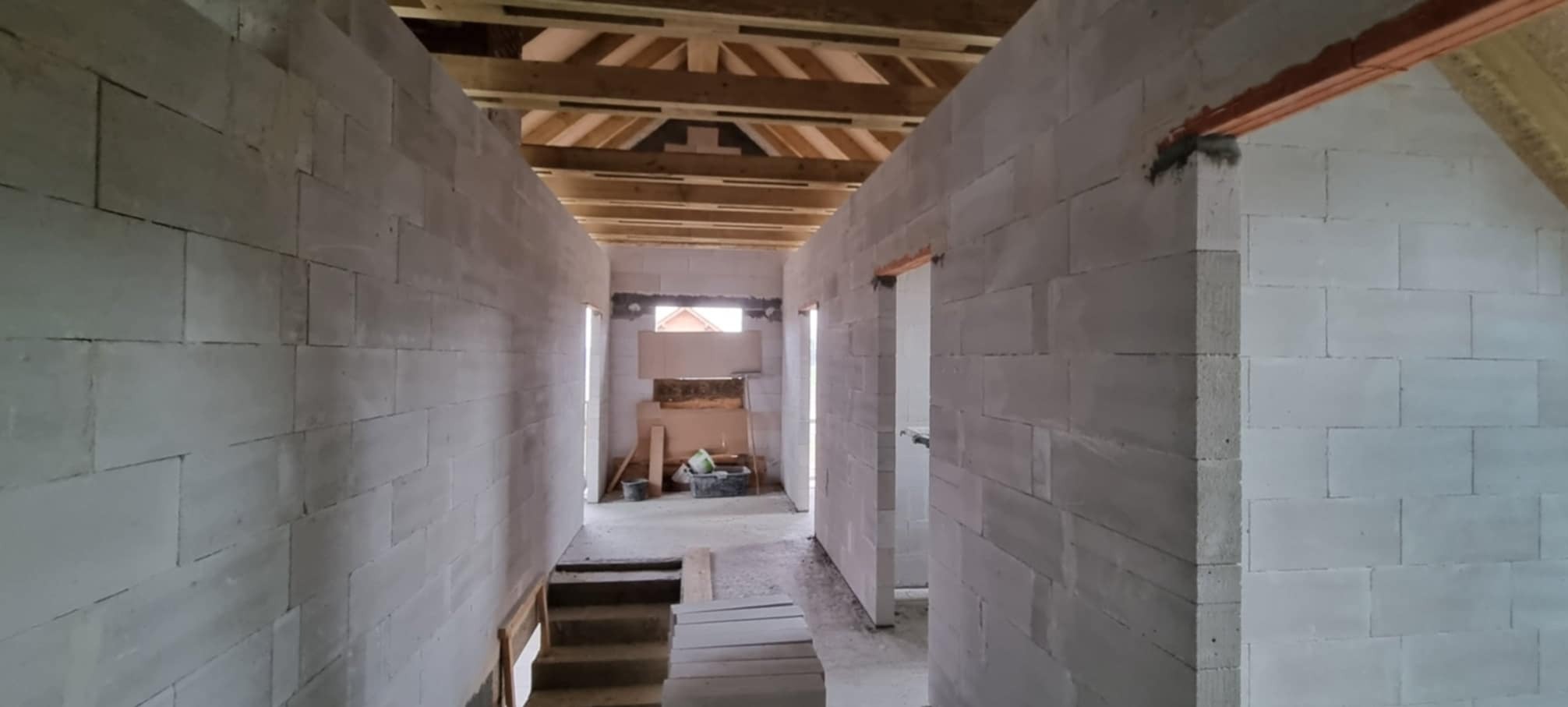 Wnętrze domu w trakcie budowy: ściany z białych bloczków, drewniany strop, widoczne przygotowane otwory na drzwi, częściowo zrobione schody, w tle narzędzia i materiały budowlane.