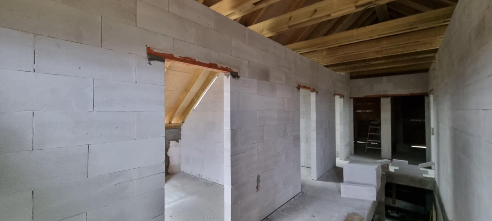Surowe wnętrze domu w trakcie budowy: ściany z bloczków, drewniana konstrukcja dachu widoczna przez otwory drzwiowe, drabina w głębi pomieszczenia.