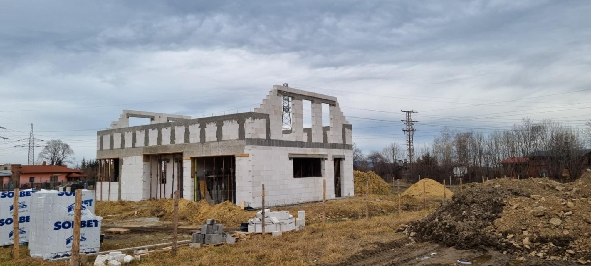 Niedokończony budynek jednorodzinny z białych bloczków, widoczne pakiety bloczków Solbet, teren budowy z piaskiem i ziemią, pochmurne niebo w tle.