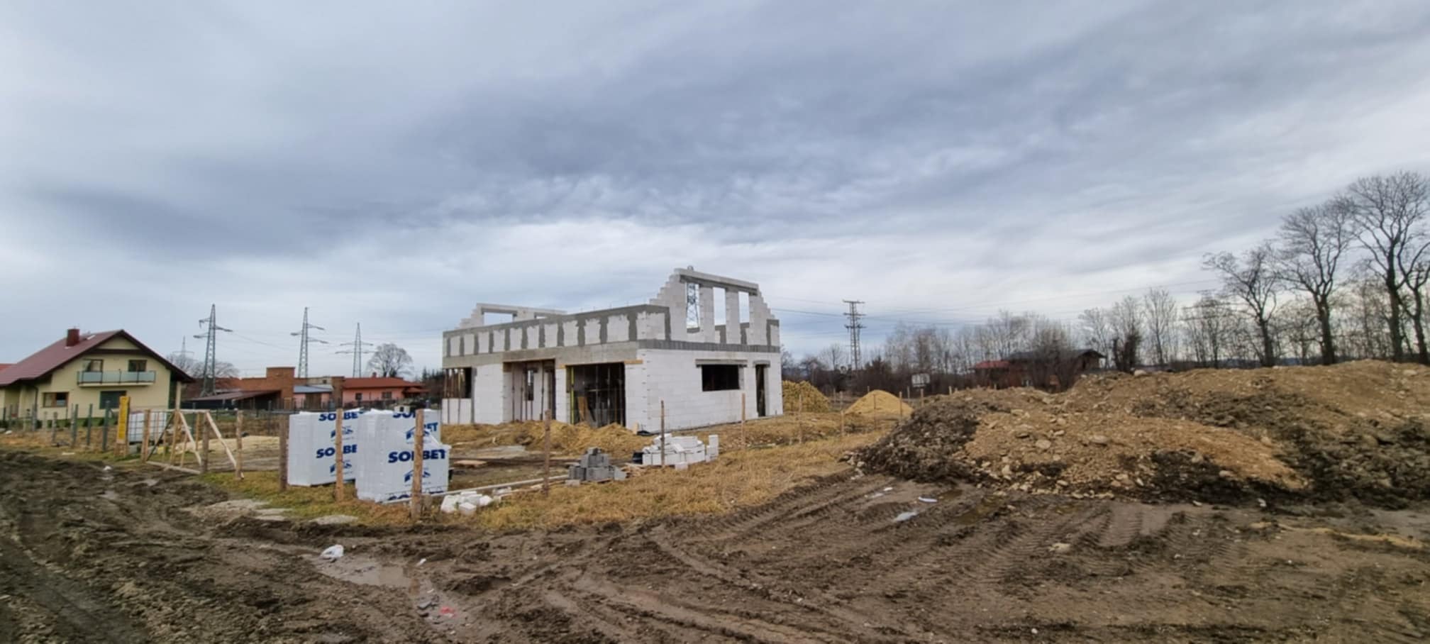 Stan surowy otwarty domu jednorodzinnego z betonu komórkowego, widoczne palety z bloczkami Sobet, w tle zabudowa jednorodzinna i pochmurne niebo.