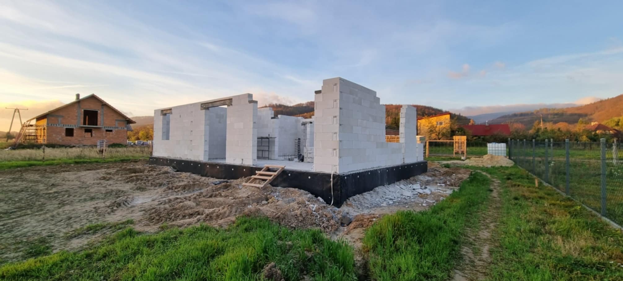 Surowy stan ścian domu z betonu komórkowego Ytong na fundamencie z czarną izolacją, widok z zewnątrz w krajobrazie z wzgórzami w tle, drewniane schodki do wejścia.