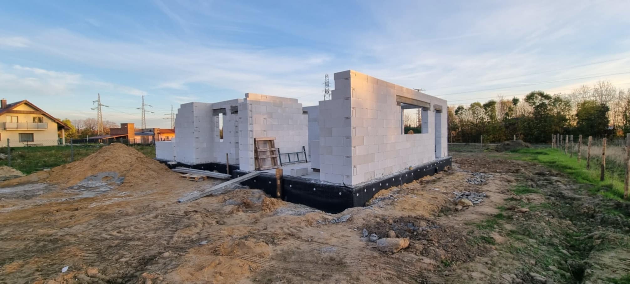 Wznoszone mury domu z białych bloczków, widoczne okna, izolacja fundamentów, ziemia i drewniane elementy budowlane na placu budowy w słoneczny dzień.