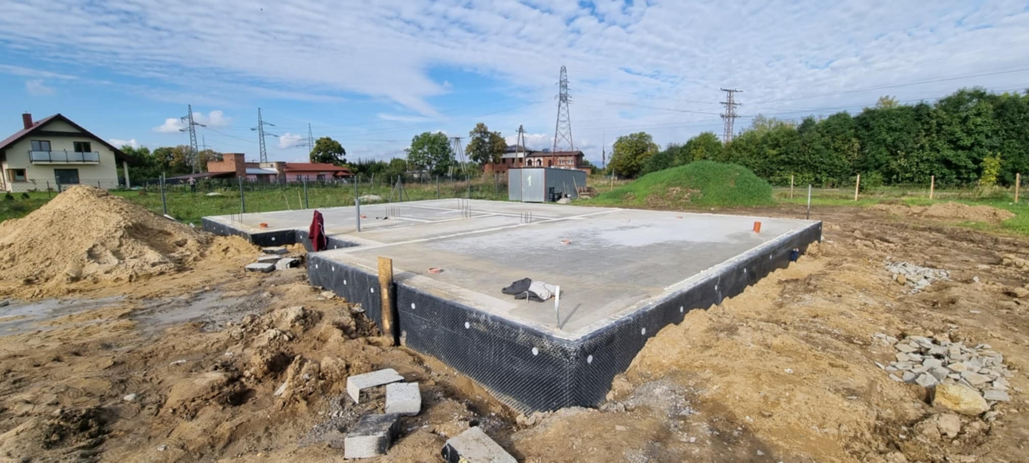 Wylany betonowy fundament z izolacją pionową w postaci czarnej folii kubełkowej, widoczne wystające pręty zbrojeniowe, otoczenie placu budowy z ziemią i gruzem, w tle dom mieszkalny i słupy...