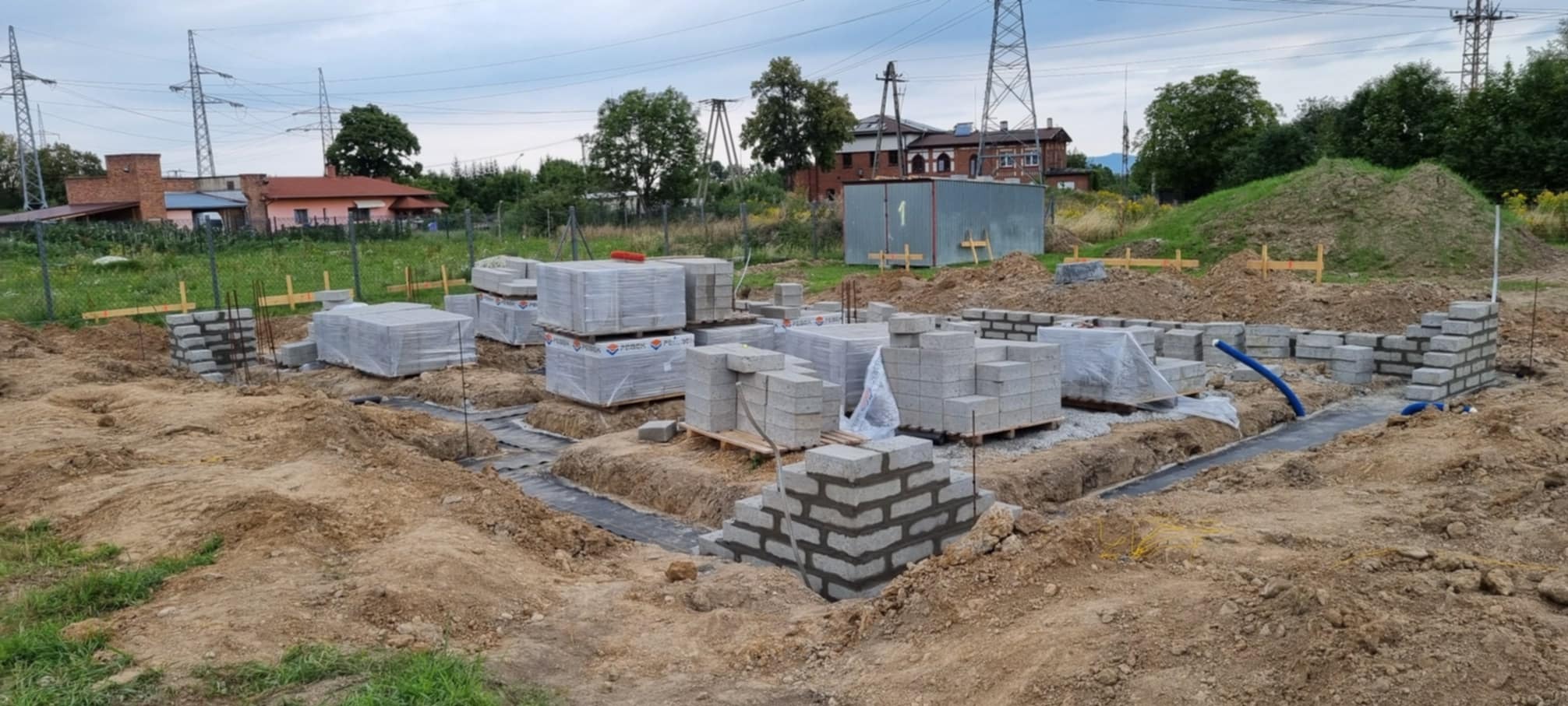 Budowa fundamentów: częściowo wzniesione ściany z bloczków betonowych, palety z zapakowanymi bloczkami, widoczne wykopy i hydroizolacja, w tle budynki i słupy wysokiego napięcia.