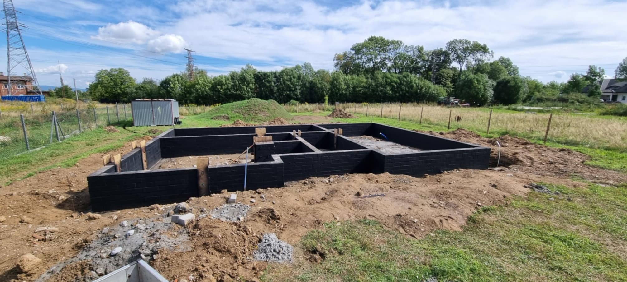 Zalane, czarne fundamenty domu jednorodzinnego na działce, widoczne drewniane szalunki i wystające rury instalacyjne, w tle słup wysokiego napięcia i zielone drzewa.