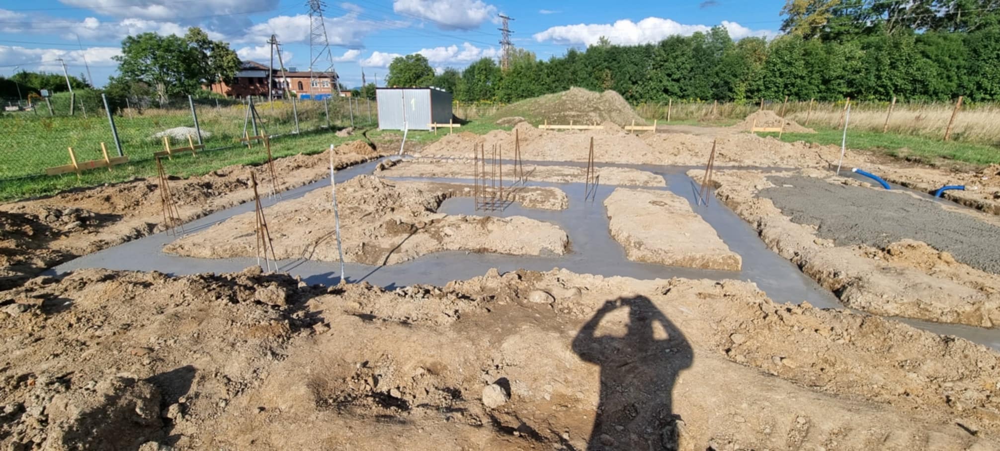Zalane betonem wykopy pod fundamenty domu jednorodzinnego z wystającymi prętami zbrojeniowymi, widok na działkę z barakiem i drzewami w tle, cień osoby robiącej zdjęcie.