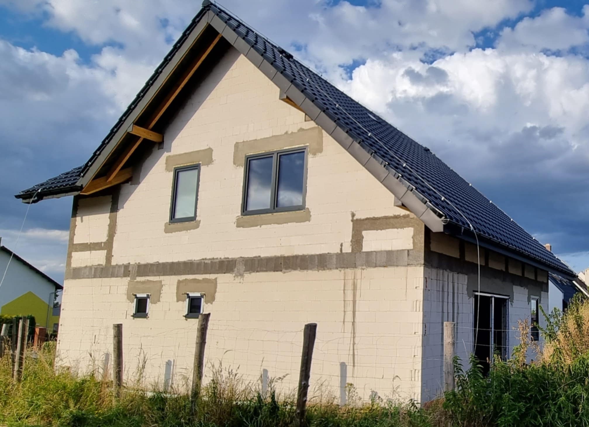 Surowy stan budynku jednorodzinnego z czarną dachówką i szarymi oknami, widoczne przygotowania pod elewację, zarośnięte otoczenie.