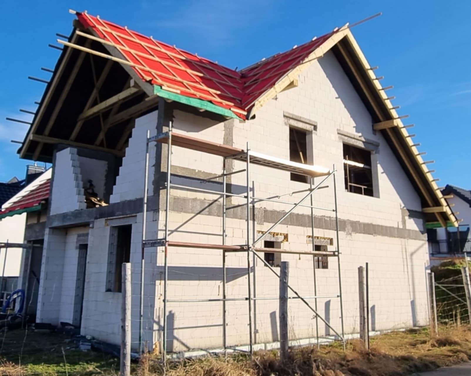 Niedokończony dwukondygnacyjny dom w trakcie budowy z częściowo ułożoną czerwoną dachówką, widoczne rusztowania i pustaki.