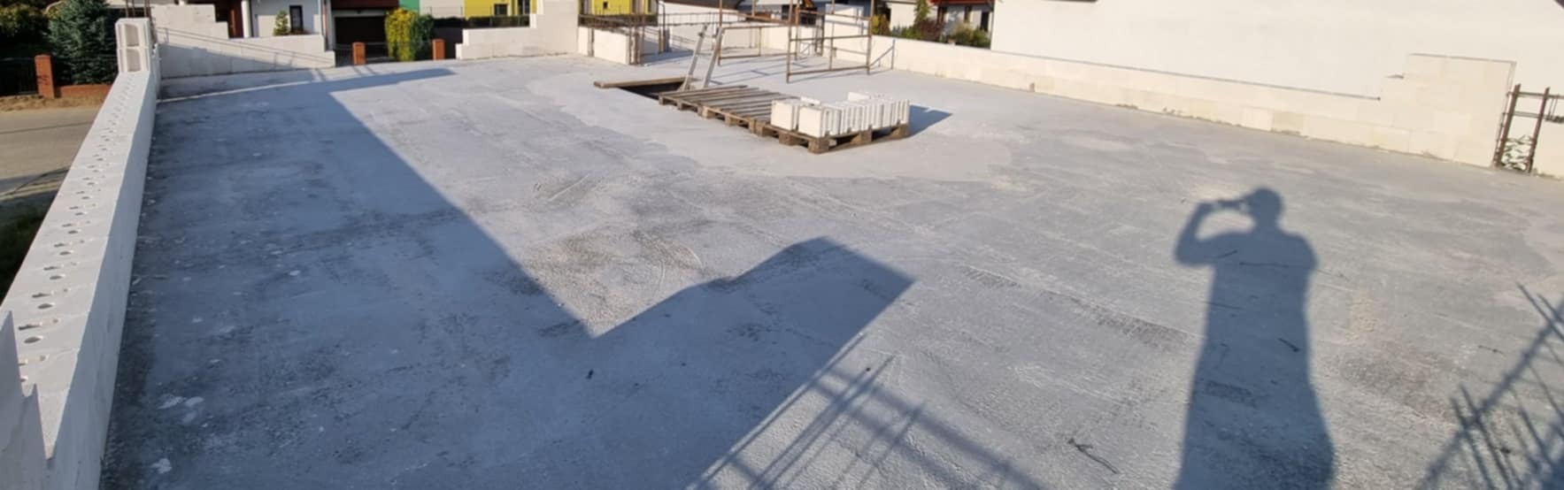 Wylana betonowa płyta fundamentowa pod budowę domu jednorodzinnego, widoczne bloczki betonowe na palecie, cień osoby robiącej zdjęcie.