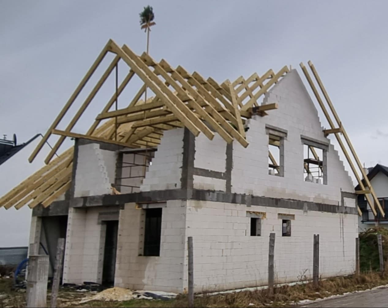 Surowy stan domu jednorodzinnego w trakcie budowy, z widoczną konstrukcją dachu z drewnianych belek i ścianami z białego bloczka, na szczycie dachu umieszczono tradycyjną wiechę.