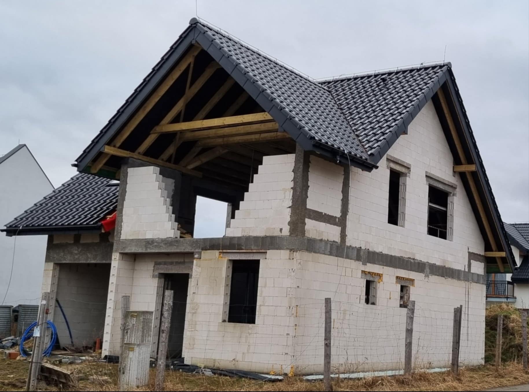 Dom w stanie surowym otwartym z czarną dachówką, widoczne drewniane elementy konstrukcji dachu i białe bloczki ścian, okna bez ram, teren wokół niezagospodarowany.