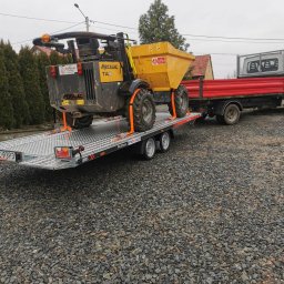 JAKUB SZCZEPANIK PROTAL - Żółta wywrotka Mecalac TA3, zabezpieczona pomarańczowymi pasami, na przyczepie platformowej. Za nią czerwona ciężarówka. W tle domy i pochmurne niebo.