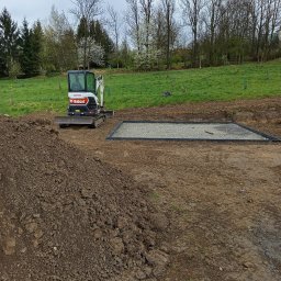 JAKUB SZCZEPANIK PROTAL - Mini koparka Bobcat na placu budowy obok przygotowanego podłoża z kamienia. W tle zielone drzewa i trawa.