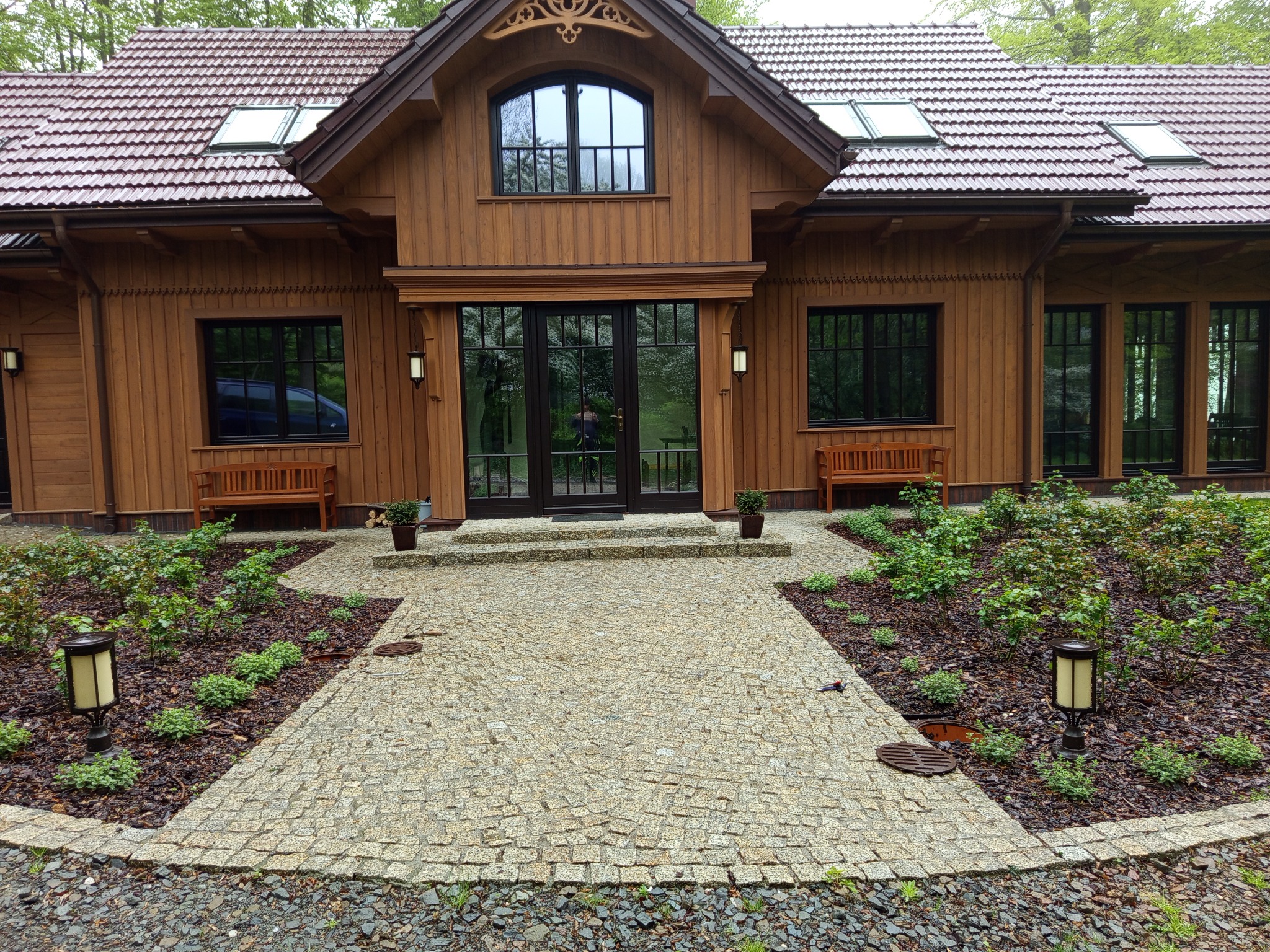 Kremowa kostka brukowa tworzy ścieżkę do drewnianego domu z ciemnymi oknami. Po bokach rabaty z roślinami i lampami. W tle zielone drzewa.