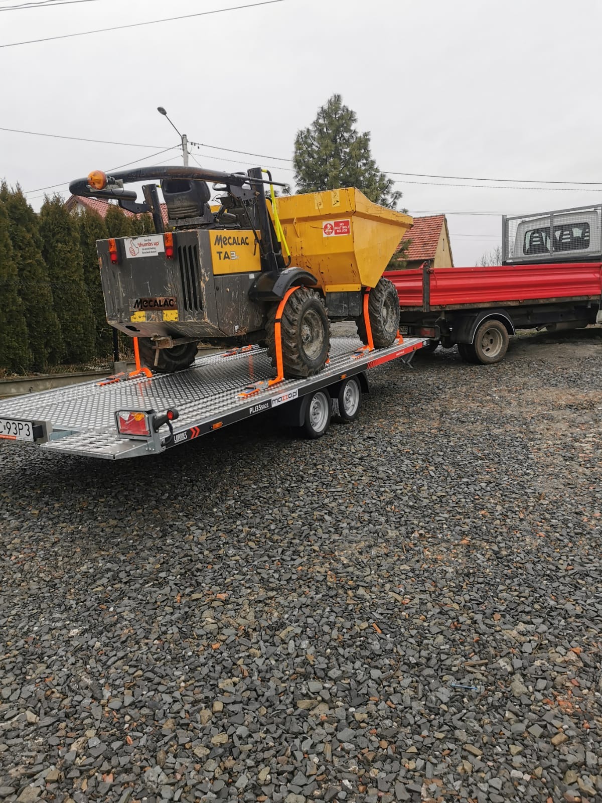 Żółta wywrotka Mecalac TA3, zabezpieczona pomarańczowymi pasami, na przyczepie platformowej. Za nią czerwona ciężarówka. W tle domy i pochmurne niebo.
