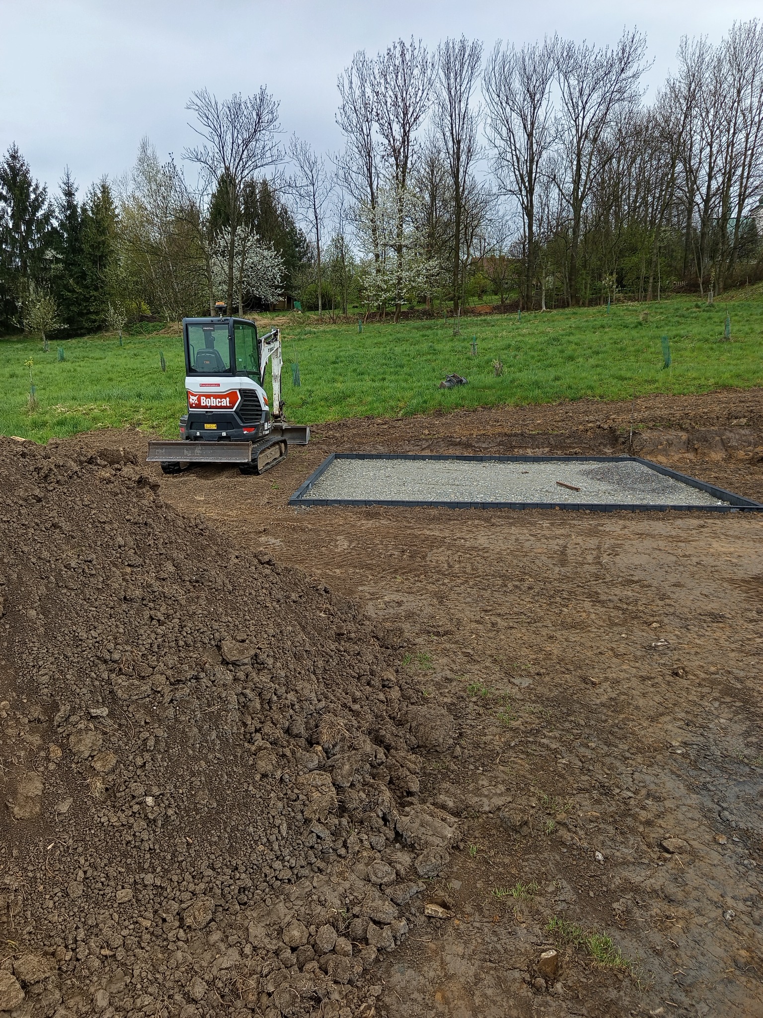 Mini koparka Bobcat na placu budowy obok przygotowanego podłoża z kamienia. W tle zielone drzewa i trawa.