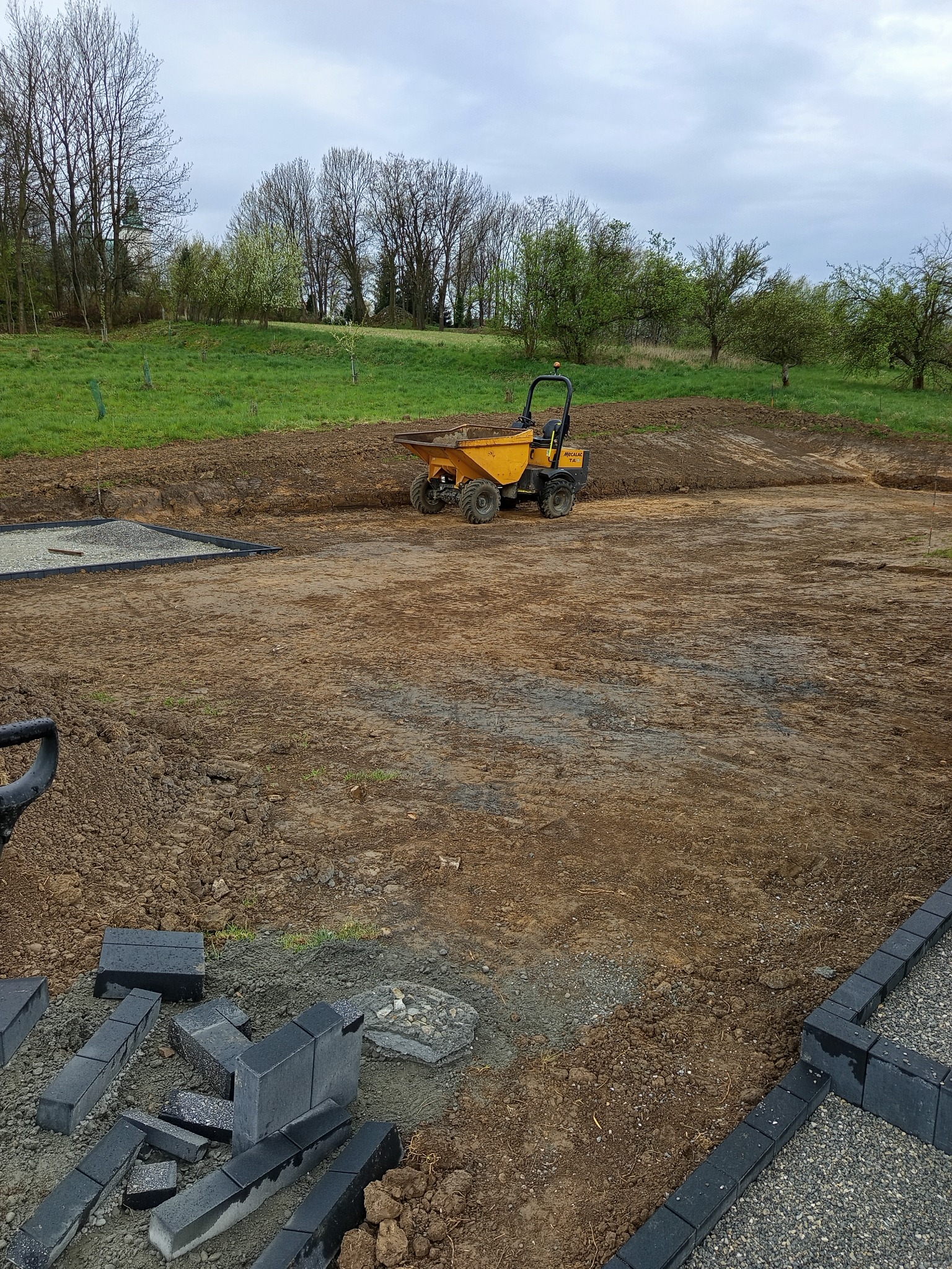 Żółta wywrotka na placu budowy, w tle zieleń i drzewa. Częściowo ułożona kostka brukowa z czarnych elementów, widoczne przygotowania do dalszych prac.